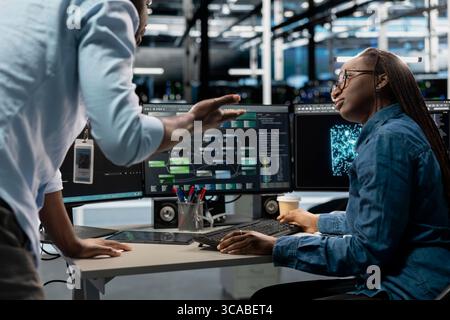 Server farm teamworking engineers doing brainstorming, looking at data analysis graph to reduce failures. Data center colleagues talking, working together using AI visualization dashboard Stock Photo