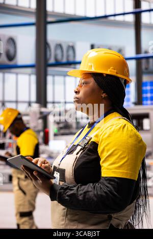 Black woman technician supervising assembly line and using tablet to ensure optimal performance. Female engineer monitors automated factory system, supervising functionality industry 4.0 software. Stock Photo