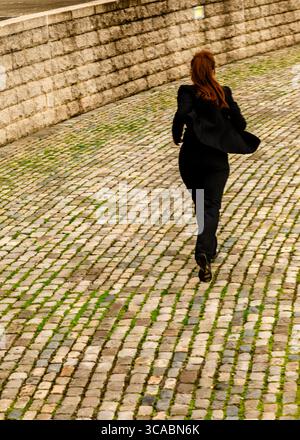 A woman in a black suit runs down a cobblestone street. Stock Photo