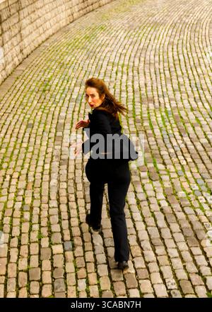 A woman in a black suit runs down a cobblestone path, looking back over her shoulder. Stock Photo