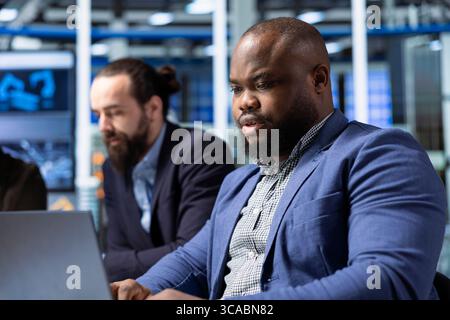 Factory engineering specialist monitoring system performance on laptop next to colleagues. African american industrial plant employee using notebook to track energy production and efficiency Stock Photo