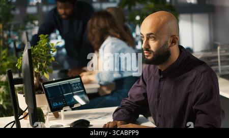 Teamworking colleagues analyzing data charts on computer screen at desk. Team of employees completing spreadsheets on desktop computer, inputting and analyzing data, camera A Stock Photo