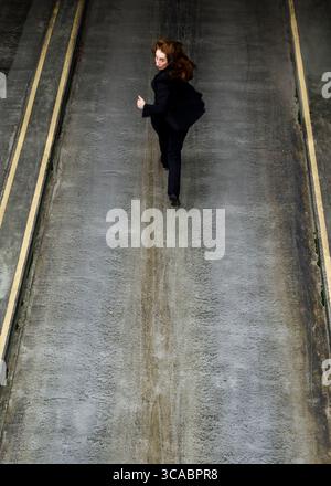 A woman in a black suit runs down a concrete path, looking back over her shoulder. Stock Photo