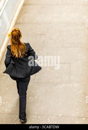 A woman in a black suit is walking on a sidewalk, possibly in the snow. Stock Photo