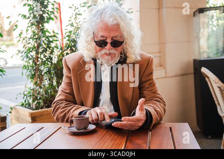 Elderly gentleman with curly hair interacting with a smartphone, sipping coffee and deeply engaged with the mobile application, showcasing a moment of Stock Photo