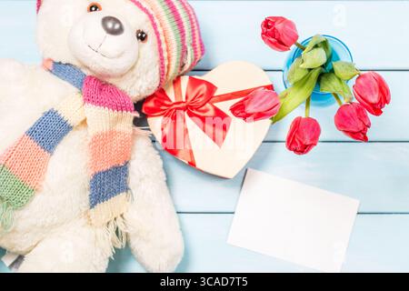 Colorful red spring tulip flowers in nice blue vase, blank photo frame and stuffed toy teddy bear with decorative heart on light wooden background as Stock Photo