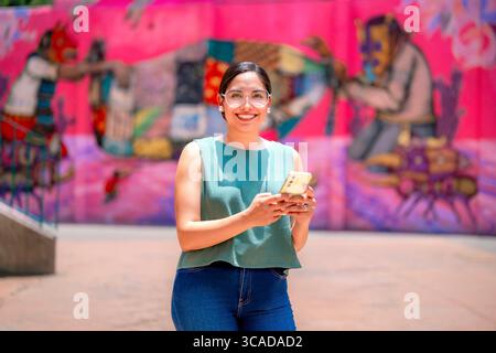 Happy young Latina woman standing in Coyoacan, Mexico City, checking her phone and smiling in front of bold graffiti at Museo Nacional de Culturas Pop Stock Photo