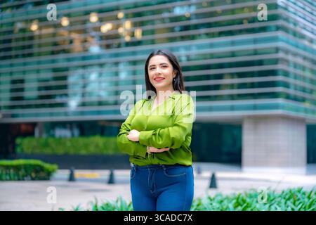 Young happy Latina business woman standing proudly with arms crossed in front of modern office building in Mexico City, wearing green blouse and blue Stock Photo