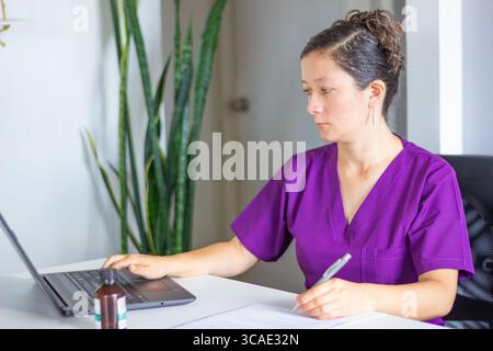 Concentrated female doctor in purple uniform using laptop and taking notes with bottle of medicine on table Stock Photo