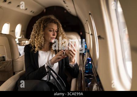 Businesswoman tapping smartphone screen in private jet cabin with blue water bottle Stock Photo