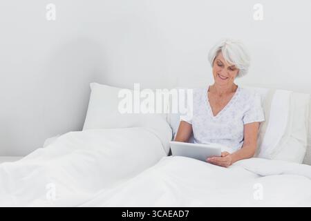 Woman sitting upright on white bedding wearing light floral nightshirt and holding silver tablet Stock Photo