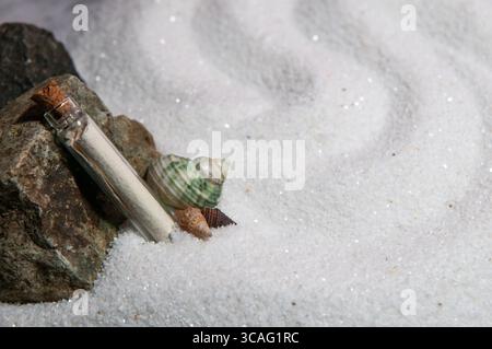 a miniature glass note in a bottle leaning against a rock with two decorative seashells on white sand. Stock Photo