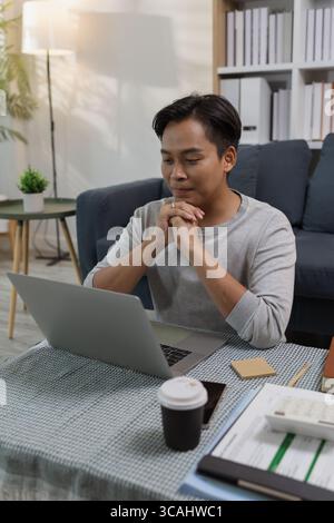 A young man sits on the floor at home, working on his laptop and thinking seriously. He is focused on his online tasks, studying, or working remotely Stock Photo