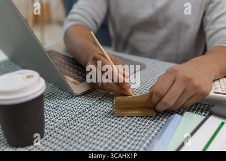A young man is working from home, taking notes with a pencil while using his laptop. There is a cup of coffee on the table, and he appears focused in Stock Photo