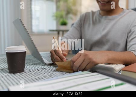 A young man is working from home, taking notes with a pencil while using his laptop. There is a cup of coffee on the table, and he appears focused in Stock Photo