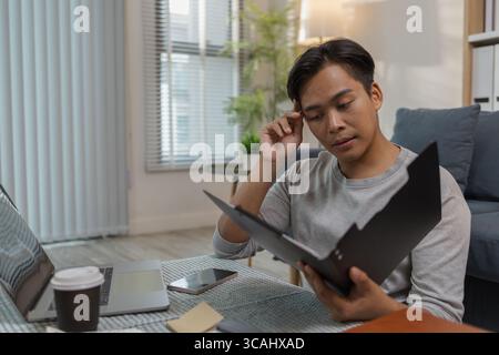 A young man is sitting on the floor at home, checking documents while working with his laptop. He appears focused and engaged, managing his tasks in a Stock Photo
