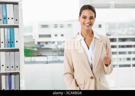 Businesswoman in suit giving thumbs-up in office by binder shelves with skyline view, copy space Stock Photo