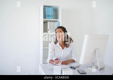 Woman sitting at office desk wearing headset, smiling and writing paperwork by monitor and binders Stock Photo