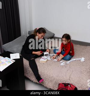 A mother and daughter are crafting on a comfy bed. The mom cuts paper with scissors, while the daughter arranges the cutouts. They enjoy a creative ac Stock Photo