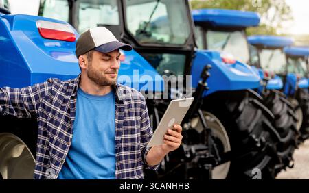Smiling man in casual clothes using digital tablet near tractors at agricultural dealership Stock Photo