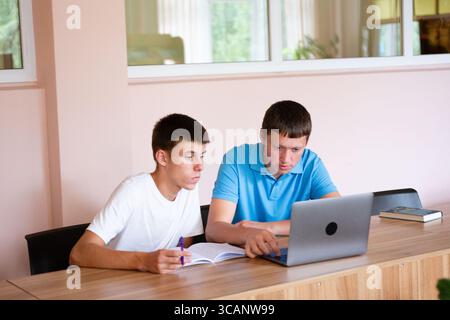 Two students are seen working together at a desk. One student is using a laptop while the other takes notes in a notebook. The setting looks like a st Stock Photo