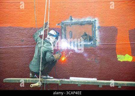 Dhaka, Bangladesh - 05 November 2021: View of a welder in protective gear skillfully fusing metal on a vibrant red and orange ship hull, sparks illuminating the scene. Stock Photo