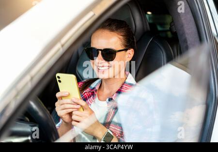Urban woman sitting in driver’s seat and using smartphone inside car Stock Photo