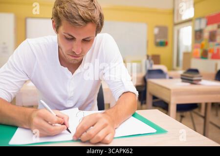 Male student leaning forward writing in binder on green folder at classroom desk under whiteboard Stock Photo