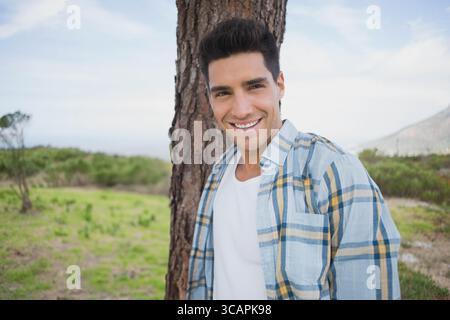 Man in his twenties leaning against tree trunk in grassy clearing with rolling hills Stock Photo