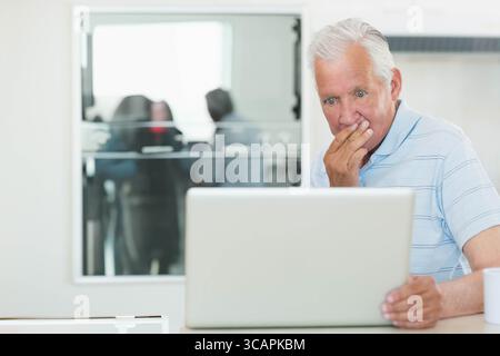 Senior man reacting with surprise at office desk examining laptop and coffee mug by glass partition Stock Photo