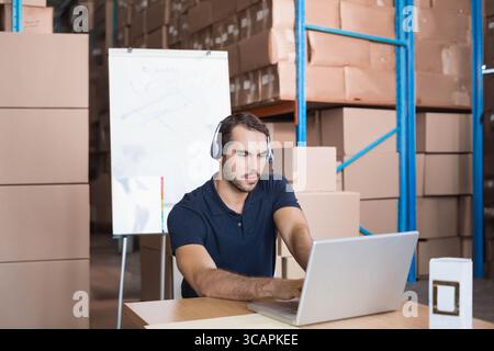 Adult male worker sitting at wooden desk in warehouse wearing headset using laptop near whiteboard Stock Photo