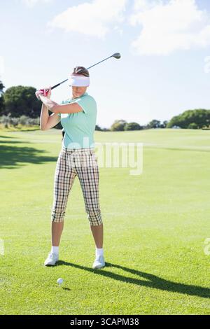 Golfer gripping club, aiming swing at golf ball teed on sunlit fairway bordered by leafy trees Stock Photo