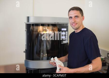 Man standing beside industrial 3D printer in workshop, holding white plastic prototype, copy space Stock Photo