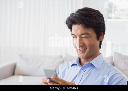 Japanese man in shirt using smartphone on white sofa, neutral cushions in living room, copy space Stock Photo