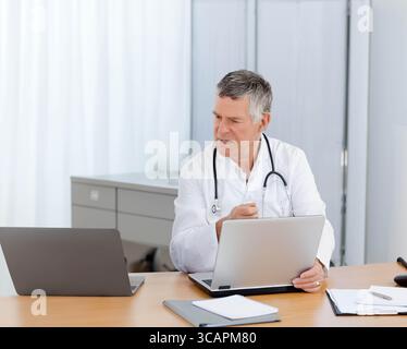 Senior male physician reviewing documents on laptop while sitting in clinic office with stethoscope Stock Photo