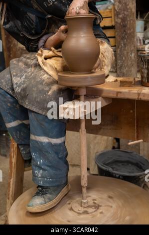 A skilled potter shapes a clay vase on a traditional foot-powered wheel in a rustic workshop. The artisan’s hands, covered in wet clay, carefully refi Stock Photo