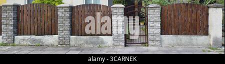 The simple fence of the old no name European house is made of vertical brown thin pine boards. Columns from a decorative white brick. Sunny spring day Stock Photo