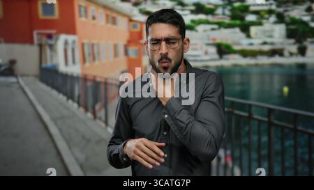 Young man in glasses and gray shirt stands on promenade near seaside holding throat with distressed expression against blurred beach backdrop Stock Photo