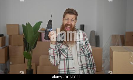Bearded man holds cordless drill and smiles inside building stacked with cardboard moving boxes in new home; excitement achievement. Stock Photo