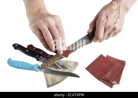 Old elderly master tries to sharpen rusty kitchen knives on his desktop. Sandpaper and grindstone are used. Isolated on white with patch Stock Photo