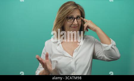 Middle aged caucasian woman wearing eyeglasses and white shirt points finger to temple in teal studio; thoughtfulness. Stock Photo