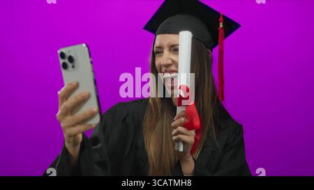 Young hispanic woman wearing mortarboard holds diploma and smartphone for selfie in studio; achievement pride. Stock Photo