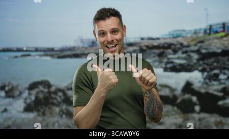 Young man with tattoos and green shirt smiles and points thumbs backwards at a rocky seaside location with a calm ocean and cloudy sky in the backgrou Stock Photo
