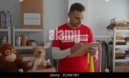 Young man wearing red volunteer shirt checking items on clipboard in charity donation room filled with clothes and toys. Stock Photo