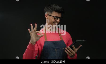 Young man with glasses and apron interacts with tablet against dark background showcasing modern technology and casual fashion style mood highlighting Stock Photo