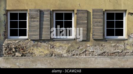 Three plastic white windows with opened wooden blings in the yellow plastered wall of a ruined old brick house. Glass isolated on black with patch. Pa Stock Photo