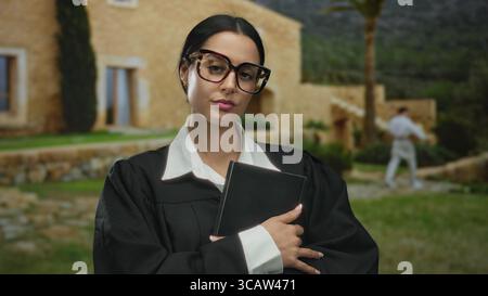 Hispanic woman in judge robe outdoors holding book in front of rustic building on a cloudy day with mountains in the background. Stock Photo