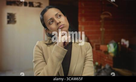 Young woman in thoughtful pose on construction site wearing tan blazer indoors conveying contemplation determination and planning within industrial se Stock Photo