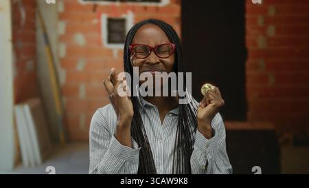 Woman holding coin with fingers crossed, african american, standing at indoor construction site, showcasing crypto enthusiasm and focusing on blockcha Stock Photo