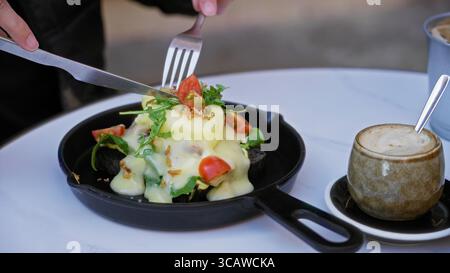 Person enjoying eggs benedict on a table with cappuccino in a sunny cafeteria showcasing vibrant salad and creamy sauce. Stock Photo